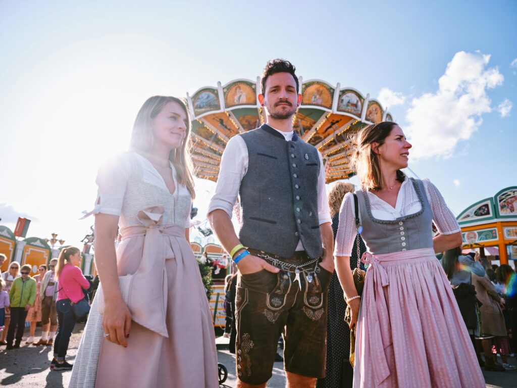 A man and to women in traditional German dress outside in front of a carnival ride at Oktoberfest in Munich, Germany