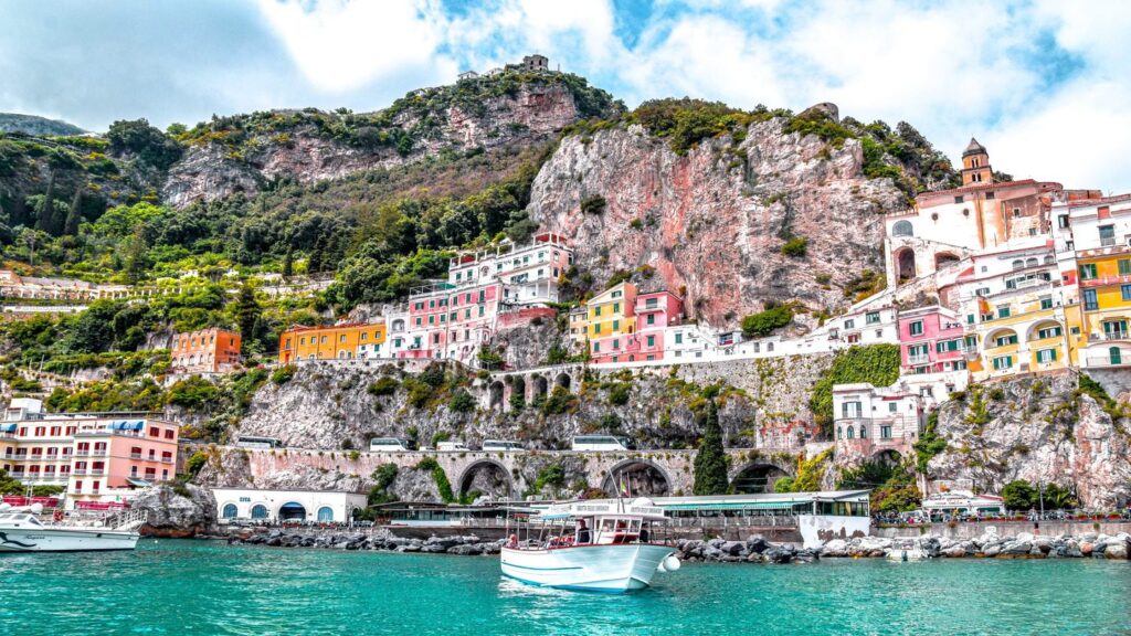 Colorful buildings nestled on rugged cliffs above a turquoise sea with a white boat floating near the shore, under a cloudy sky in Italy.