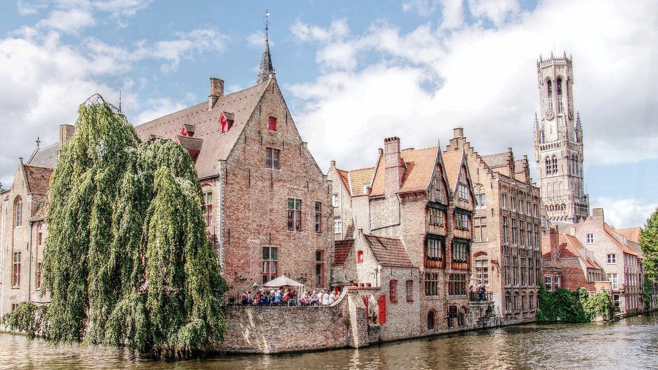 Historic brick buildings and a tall, slender tower beside a tranquil canal, populated with lush trees and a clear sky, in Bruges, Belgium, perfect for your winter travel bucket list.
