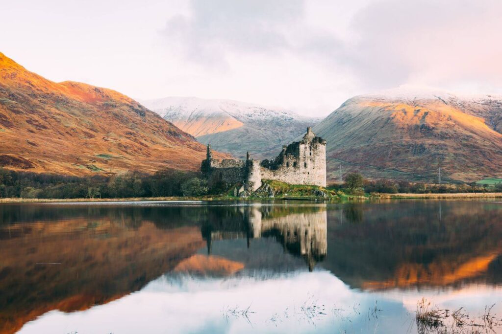 A serene heritage destination featuring an ancient castle mirrored in a calm lake, flanked by autumnal hills and snow-capped mountains.