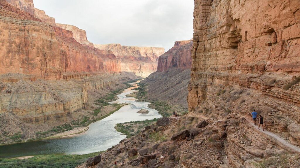 Two people are walking down the Grand Canyon near a river.