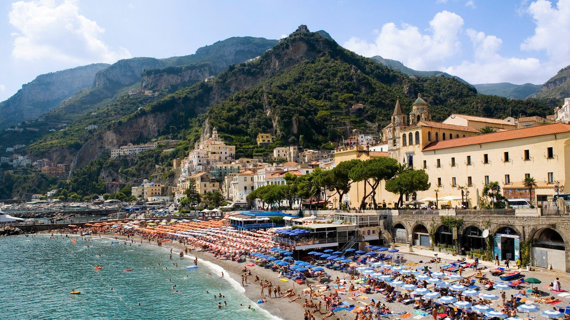A beach and sea front with jagged green hills in the background and turquoise water