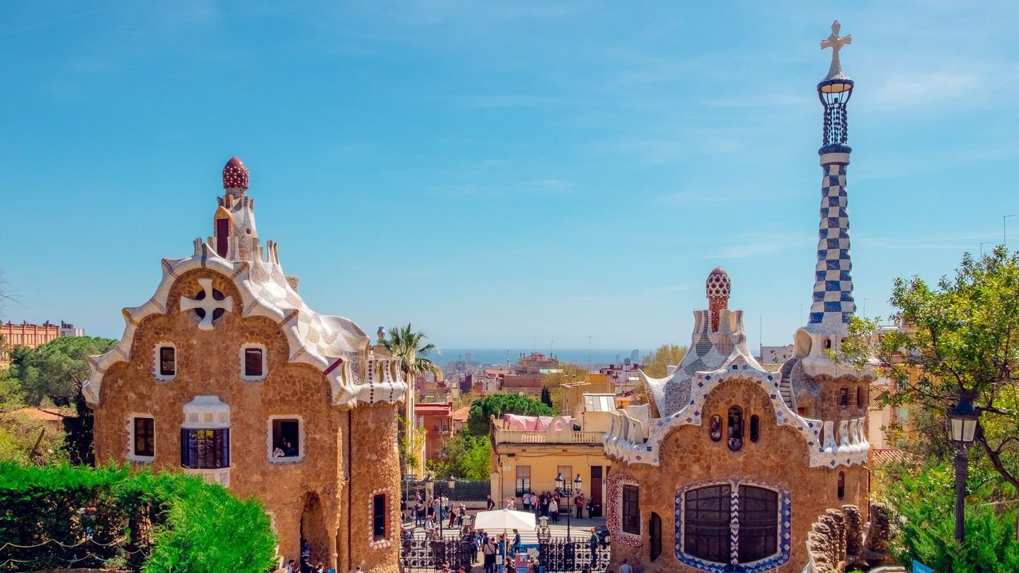 Two distinctive buildings with unique, mosaic-tiled roofs at the entrance of Park Güell in Barcelona, with visitors enjoying spring tours and a clear blue sky.
