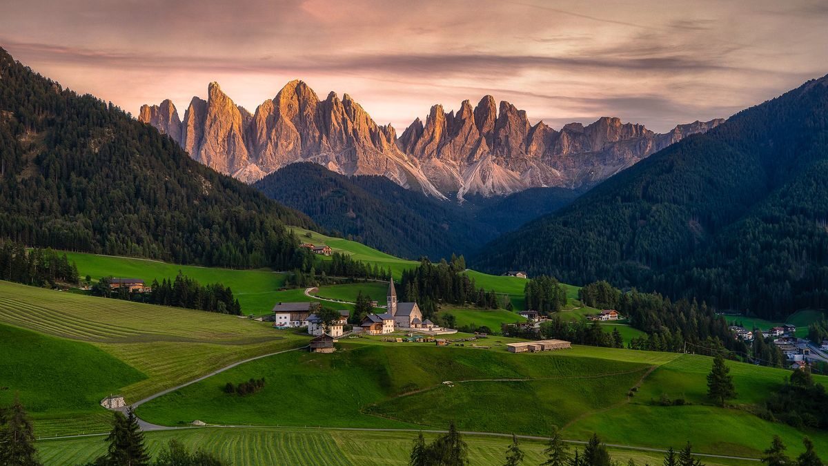 Sunset over a lush valley with a small village, featuring prominent, jagged mountain peaks in the background during a Trafalgar trip.