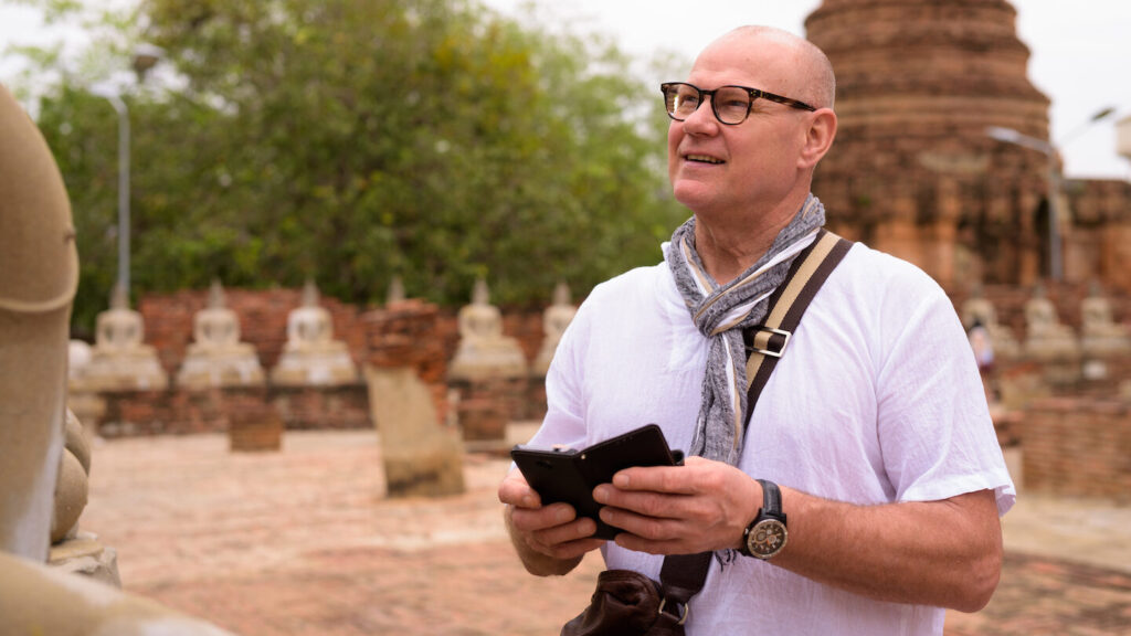 A bespectacular man holding a smartphone smiles while standing at an ancient temple site with buddha statues in the background, perfectly illustrating what to bring to Asia.