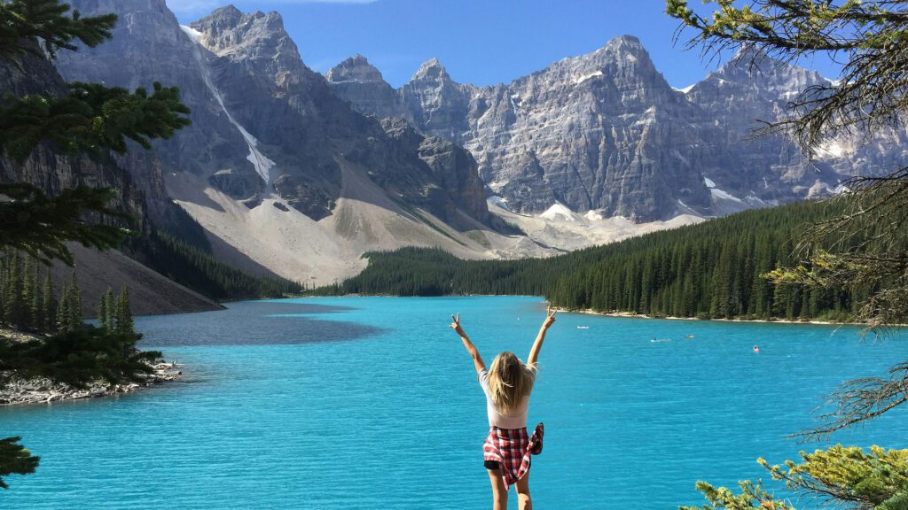 Woman standing with arms raised in the air overlooking a bright blue lake with mountains and forested hills in the background